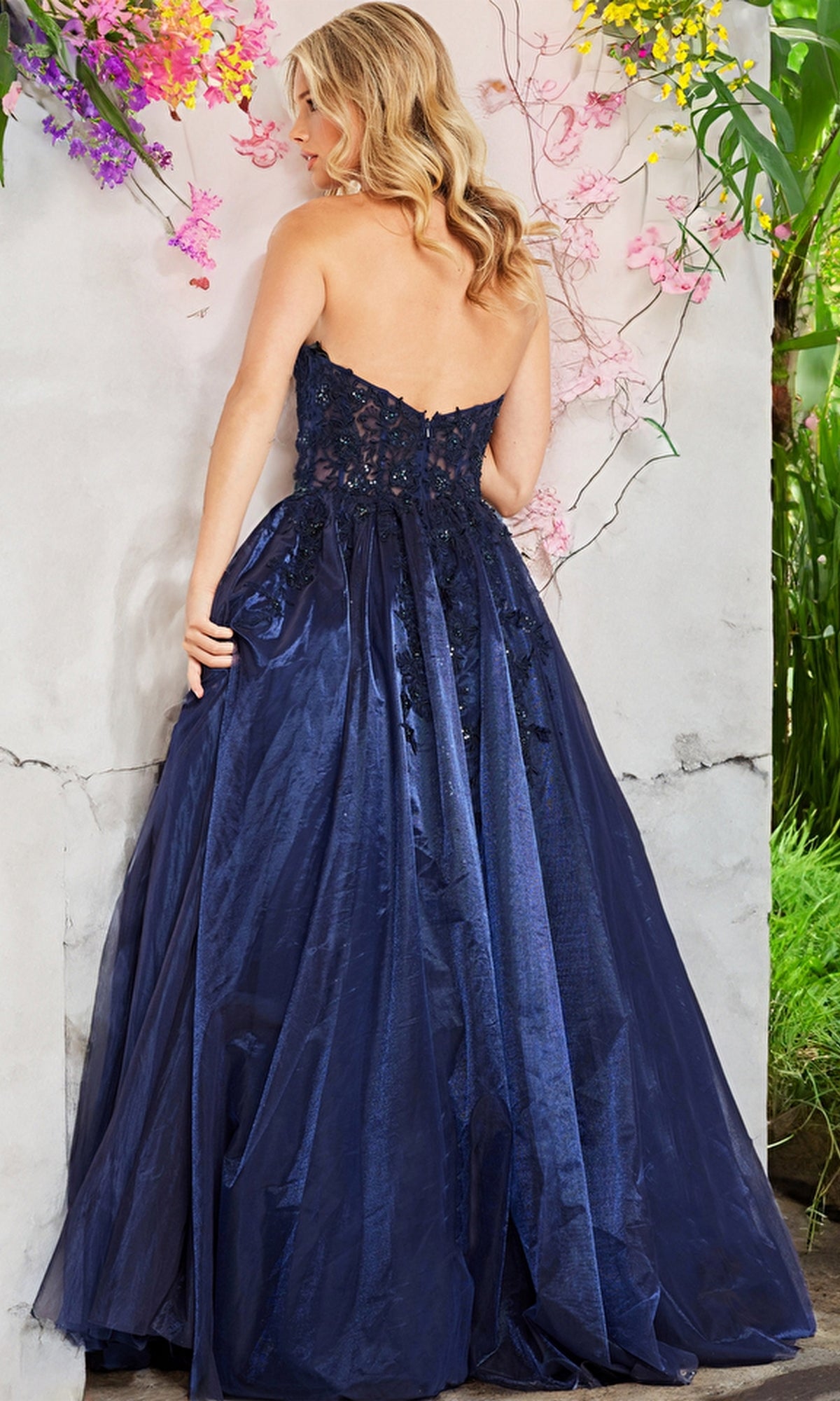 A woman with wavy blonde hair wears the Jovani 00915 Strapless Long Ball Gown, featuring a fitted bodice and full skirt in navy blue, as she stands before a flower-decorated wall.